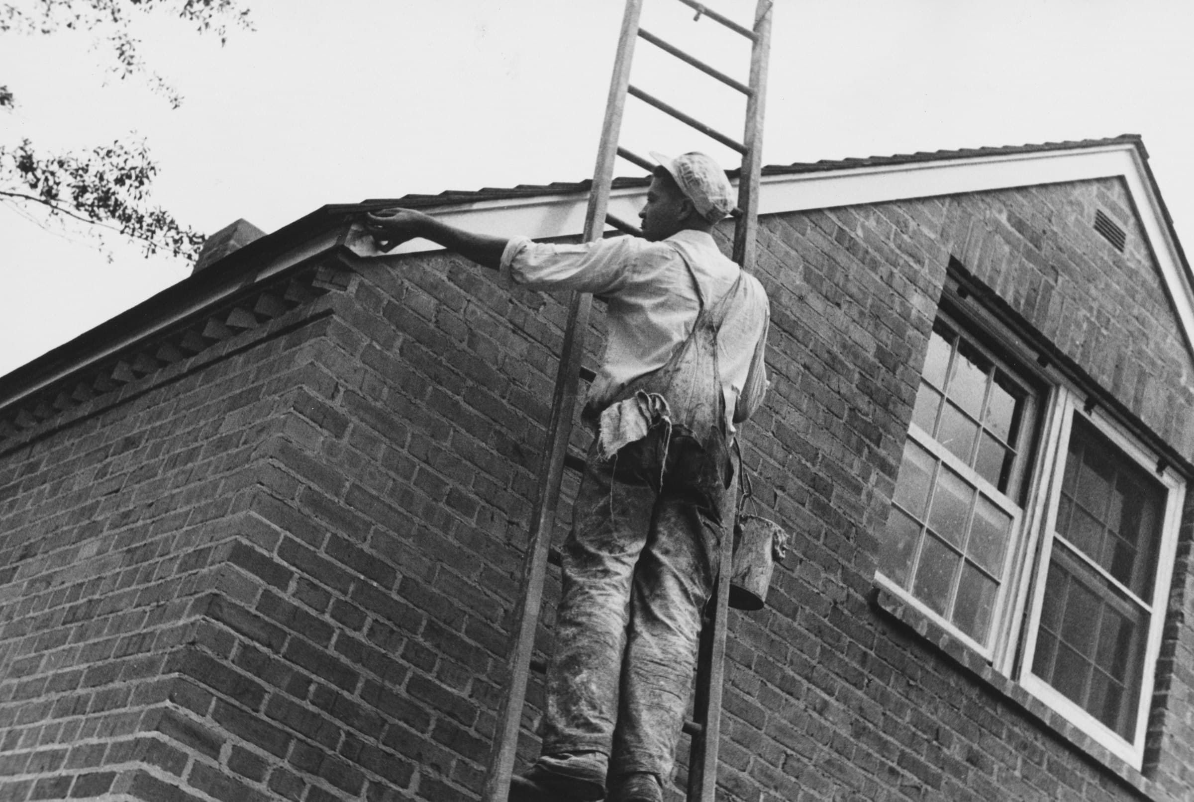 A vintage-style photograph of a painter on a ladder finishing trim on a brick house