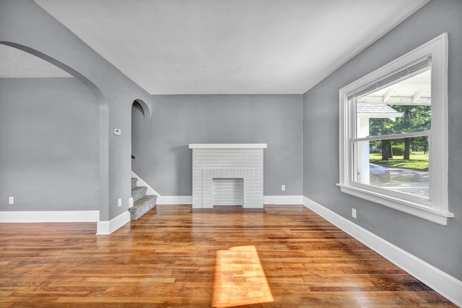 An empty room with grey walls, hardwood floors, and a white brick fireplace awaiting a fresh coat
